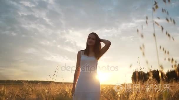 一个年轻的姑娘站在金色的田野的小麦在日落时分和引发他举起手来，慢动作