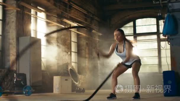 积极在健身房运动的女性在她交叉健身锻炼锻炼与战斗绳索 / 高强度间歇训练。她是肌肉和冒汗，健身房是在废弃的工厂