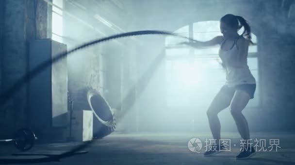 坚强的竞技女人练习用绳索作为一部分的她交叉健身健身房锻炼。她被覆盖在汗水与培训发生在废弃的工厂，改造成健身房
