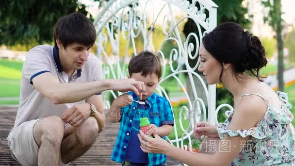 年轻的父母教他们的儿子到吹泡泡吗，妈妈抱着一罐液体，爸爸怕吹，宝宝最喜欢一个新的游戏