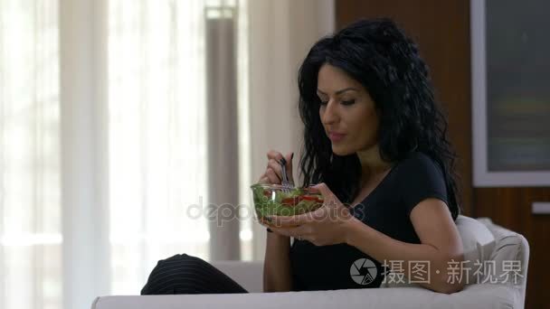 健康的生活方式，吃一碗新鲜的蔬菜沙拉在家里的沙发上坐着的女人