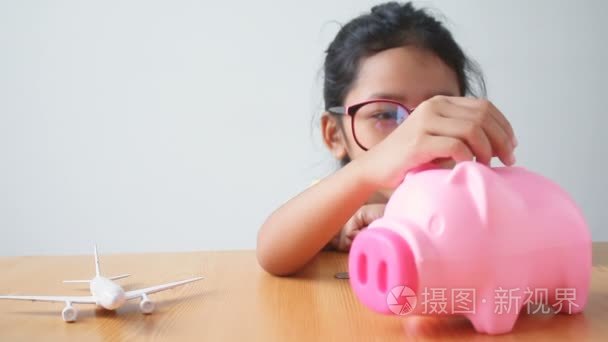 亚洲小女孩硬币投入一个粉红色的存钱罐，与空气平面隐喻为旅行和运输的概念存钱