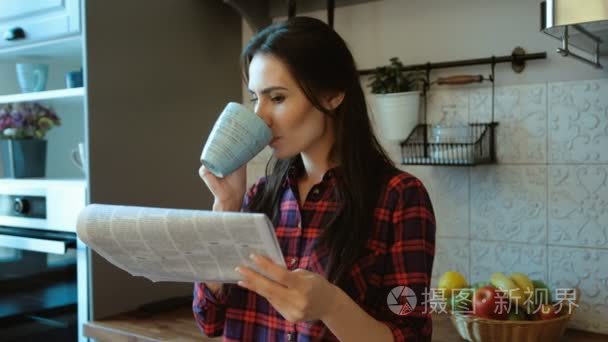 年轻漂亮的女人喝咖啡和阅读早报站在厨房里。关闭