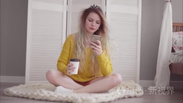 年轻漂亮的女性坐在她的卧室的地板上, 喝咖啡和使用智能手机。风吹动头发