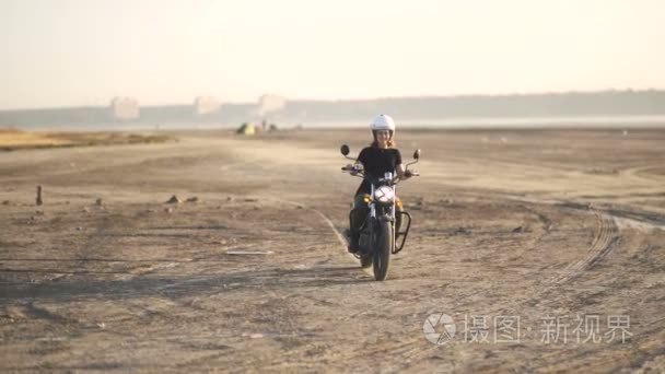 美丽的年轻女子骑旧的咖啡馆赛车摩托车在沙漠上在日出或日落时。女骑自行车的人。慢动作
