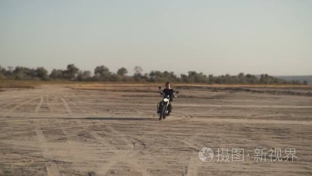 美丽的年轻女子骑旧的咖啡馆赛车摩托车在沙漠上在日出或日落时。女骑自行车的人。慢动作