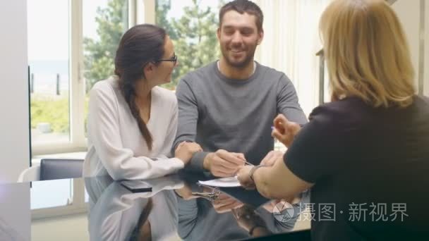 坐在桌前生活美丽 年轻夫妇签订合同，买房子，房地产经纪人给了他们家的钥匙。新婚夫妇幸福地拥抱。家是阳光和光明