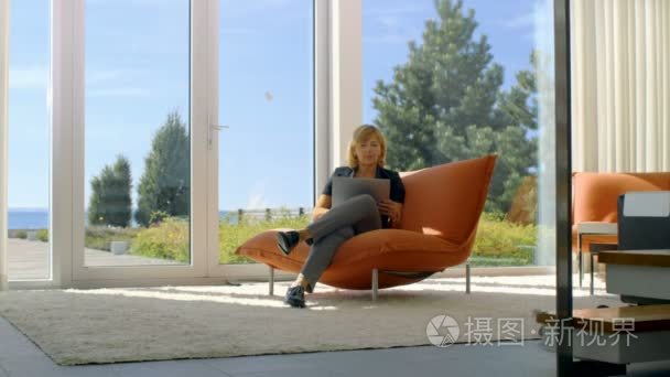 高级的女人使用笔记本电脑坐在她阳光的客厅里的沙发上。房子有地板到天花板的窗户与拥有视图。天气晴朗