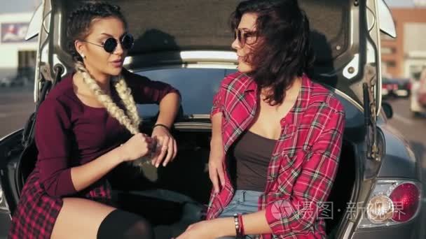 两个吸引力的年轻女性在时尚太阳镜坐在汽车后备箱在停车场的购物商场和谈话期间阳光灿烂的日子。慢动作镜头