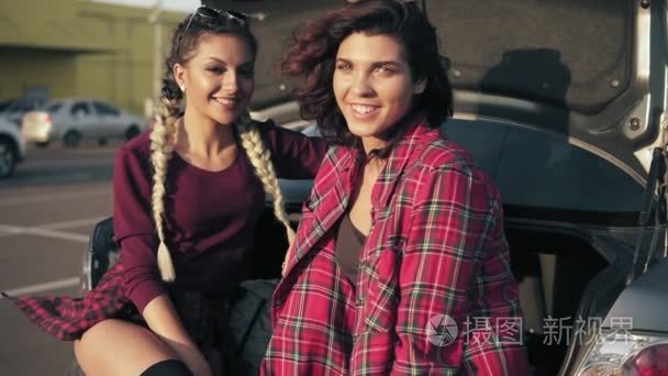 两个年轻漂亮的女性坐在停车场的汽车后备箱由购物商城在阳光明媚的日子，微笑着看着镜头。慢动作镜头
