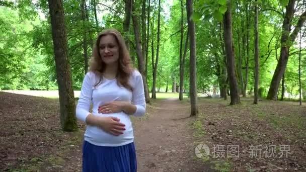 微笑的怀孕妇女独自一人走在公园和抚摸她的肚子