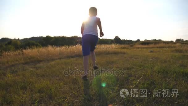 之后到阳光明媚的日子在绿色草地上的字段上运行的小男孩。慢跑在草坪户外与太阳耀斑的孩子。男性的孩子会在自然夏天草地上。慢动作后方回