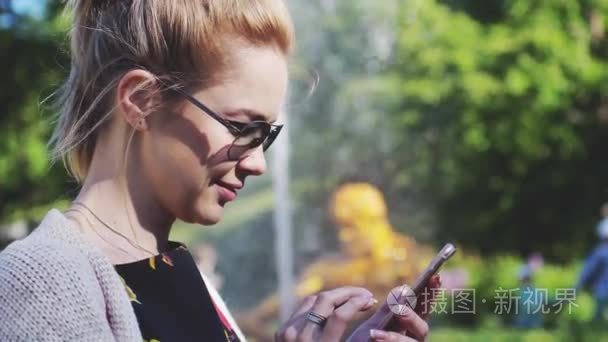 开朗的女人，戴着墨镜在公园与一部智能手机。一个女人模糊喷泉在背景上她手机写一条消息。慢动作。1920 x 1080