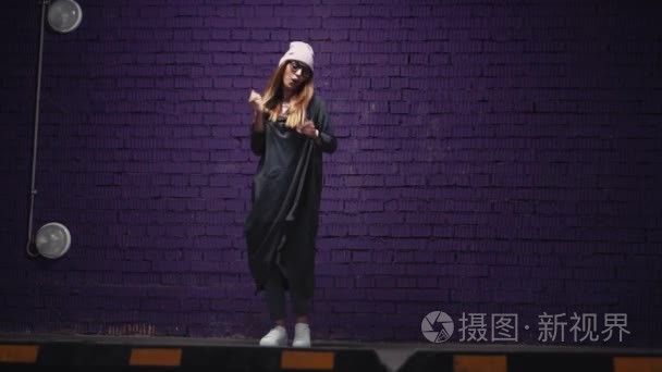 一个时尚的时髦女孩在时尚的眼镜和帽子跳舞附近的砖紫色墙的耳机。慢墨视频