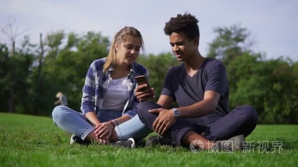混的血夫妻坐在草和选择的图片，在自己的智能手机。年轻人微笑，笑出声。可爱的朋友在公园里，坐在草地上休息享受智能手机