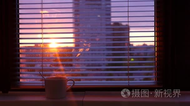 窗台上有一杯热咖啡  在开放的百叶后  你可以看到城市和夕阳光线穿透房间。slowmotion.1920x1080