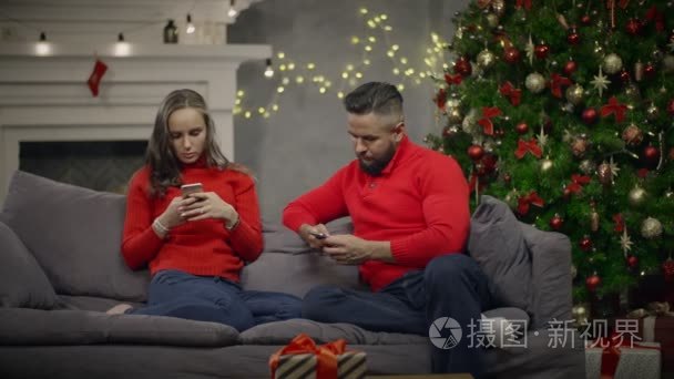 年轻夫妇坐在沙发上  在背景圣诞树上使用智能手机