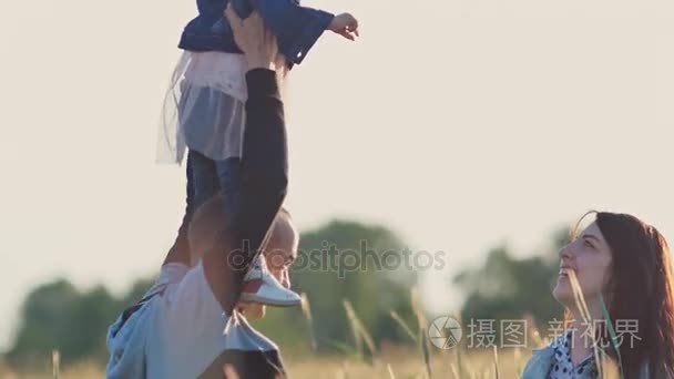 幸福的年轻家庭的三人一起在一个麦田里的绿色小穗。一个小女儿坐在教皇的肩膀上。他们一起在夏天享受大自然。父母玩