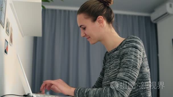 一个年轻的女人坐在桌子旁的看法  与笔记本电脑和打字文本快速查看屏幕和微笑