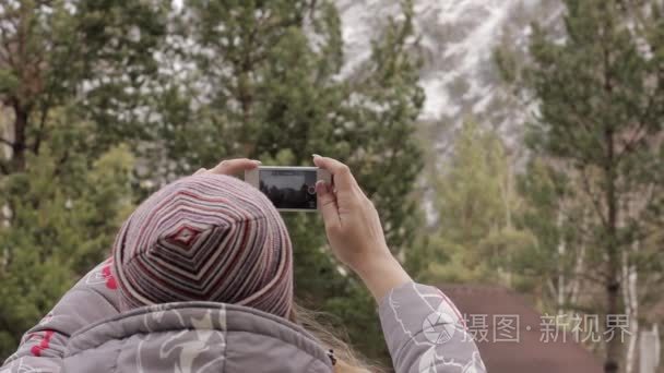 年轻的女游客在她的智能手机上拍摄山和松树