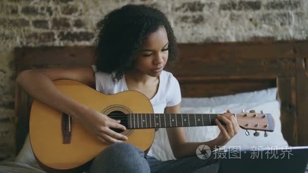 年轻的卷发混合种族女孩 concentraing 学习弹吉他使用膝上型电脑坐在家里的床上