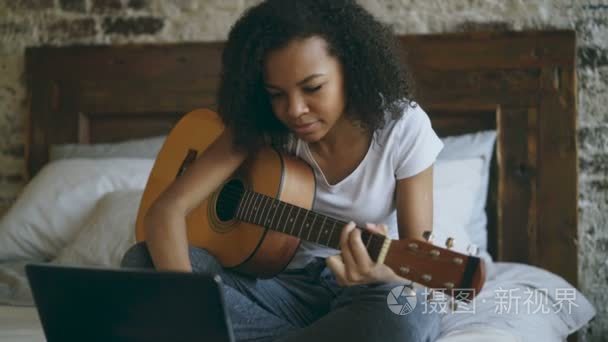 迷人的非洲裔美国少女 concentraing 学习弹吉他使用膝上型电脑坐在家里的床上