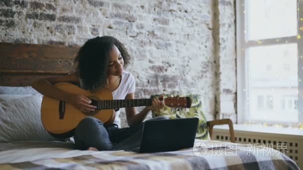 卷曲的非洲裔美国少女 concentraing 学习弹吉他使用膝上型电脑坐在家里的床上