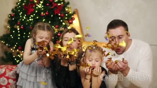 四的年轻幸福家庭坐在圣诞树旁, 吹着金色的五彩纸屑。可爱的母亲, 父亲和两个女儿一起庆祝圣诞节。Slowmotion 