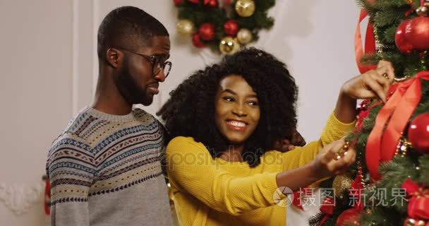 接近年轻迷人的微笑的非洲裔美国夫妇准备圣诞节和装饰圣诞树与红丝带和金色小。在客厅里肖像拍摄