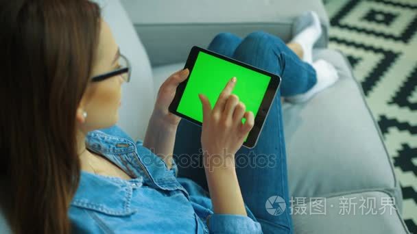 美丽的年轻女子使用黑平板电脑与绿色屏幕, 而坐在沙发上的客厅里。女人手滚动, 滑动页。色度键。顶部视图