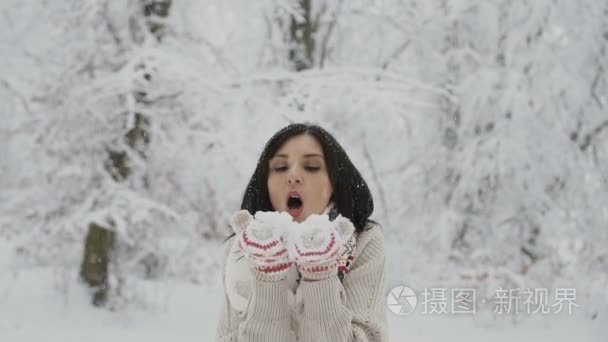 特写.冬天是年轻女子吹雪她的针织手套女孩在冬季公园的乐趣。在户外享受乐趣的美丽的少妇