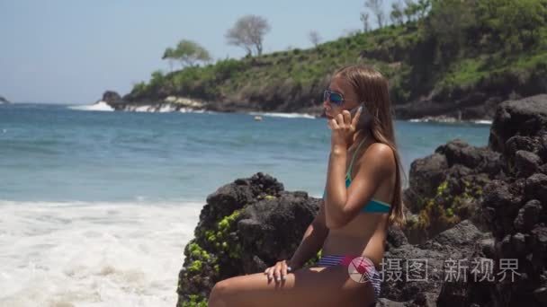 有手机的女孩坐在岩石上  看着大海。印度尼西亚  巴厘岛