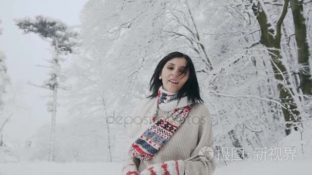 一个年轻的黑发女子的特写镜头  他在寒冷的冬日里看着镜头  穿着时髦的羊毛毛衣、暖和的围巾和针织手套在白雪覆盖的森林里漫步。