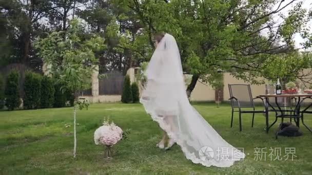 美丽的金发新娘在一个豪华的长结婚礼服漫步在绿色花园。新娘的早晨。婚礼日