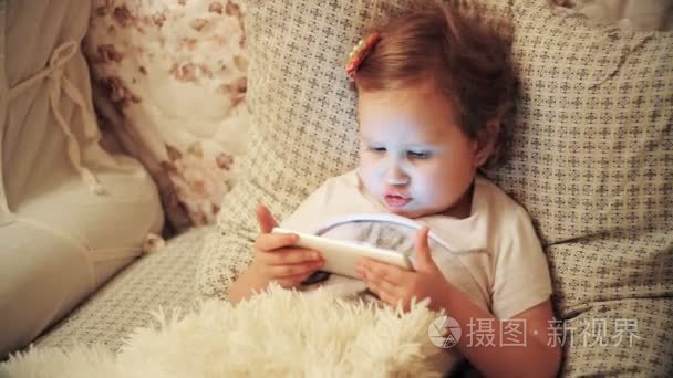 肖像特写滑稽的孩子看着手机屏幕和播放下载的应用程序在智能手机特写。一个可爱的小女孩躺在床上的毯子下的卧室和玩游戏视频