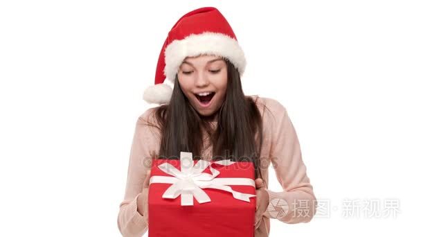 长头发的兴奋女性的肖像戴着圣诞老人的红帽被震惊地收到精美的新年礼物裹在白色背景。情感概念