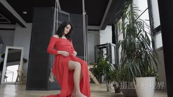 在一个绿色的花附近的时尚工作室, 一个怀孕的女人坐在秋千上轻轻地抚摸她的胃。怀孕的概念, 幸福的家庭, 不久的母亲, 男婴