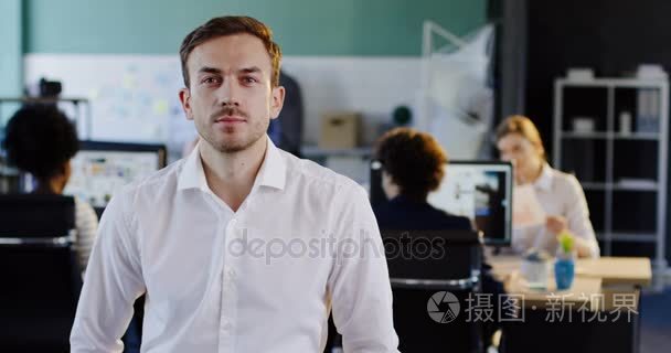 有吸引力的年轻人穿着白衬衣的画像  看着摄像头  在电脑后面的办公室里找不到工作的人。室内