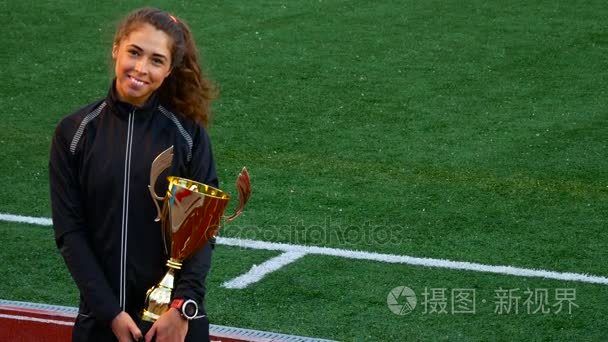 愉快的微笑的年轻妇女以胜利战利品金杯奖在橄榄球领域背景