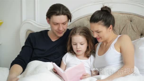 漂亮的小女孩正试图与她的父母躺在床上看书。美丽的孩子正在看书  用手指在书上显示