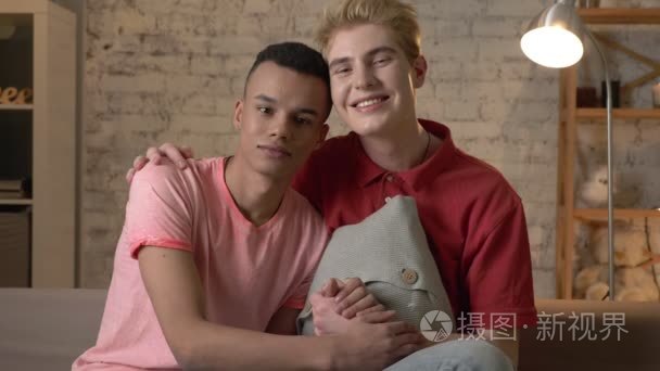 两个多民族的同性恋朋友坐在沙发上手拉手  拥抱  微笑  看着镜头。家舒适  家庭  lgbt。60 fps