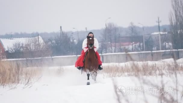 穿着红色连衣裙的年轻黑发女郎在冬天的雪覆盖的田野上飞快地奔驰在一匹马上。