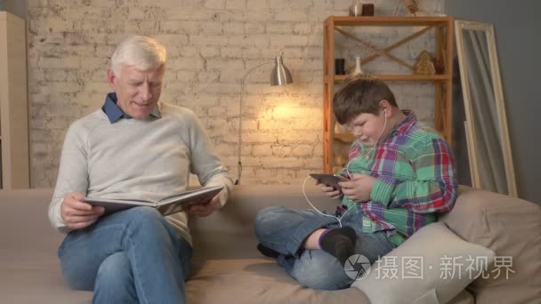 世代的区别。老人坐在沙发上看书  一个年轻的胖子用平板电脑和耳机说话。家居舒适  家庭田园风光  舒适概念 60 fps