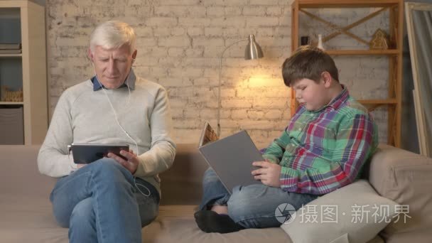 世代的区别。一个老人坐在沙发上使用平板电脑和耳机  一个年轻的胖男孩正在看书。家居舒适  家庭田园风光  舒适概念 60 fps