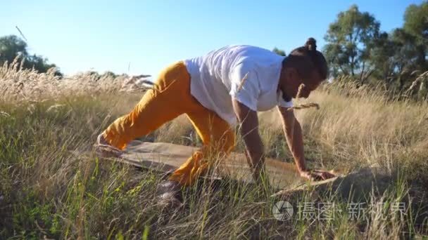 年轻人练习瑜伽动作和立场在垫子上的领域。在阳光明媚的日子  运动的家伙在草地上做运动。夏季景观背景。健康活跃的生活方式的概念。特