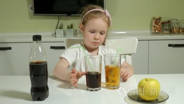 一个可爱的小女孩坐在桌子旁, 选择喝什么, 可乐或果汁, 健康生活方式的概念。