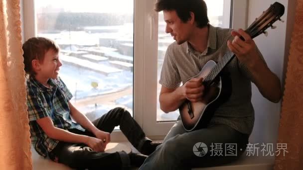 演奏乐器。爸爸正在弹吉他, 儿子在窗台上玩着铃鼓。