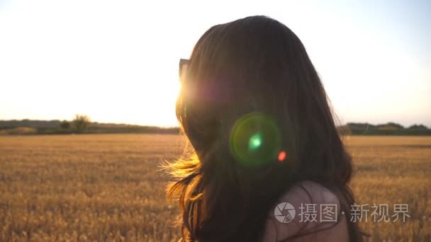 美丽的女孩正沿着麦田漫步, 背景是太阳耀斑。在日落的草地上的年轻女子的轮廓。夏天休闲在自然概念。侧面视图关闭慢动作视频