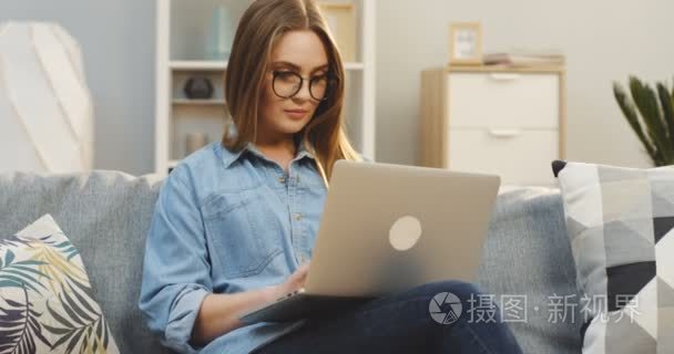 穿着牛仔裤和眼镜的漂亮年轻女子在笔记本电脑上工作, 在舒适的起居室里上网冲浪。室内