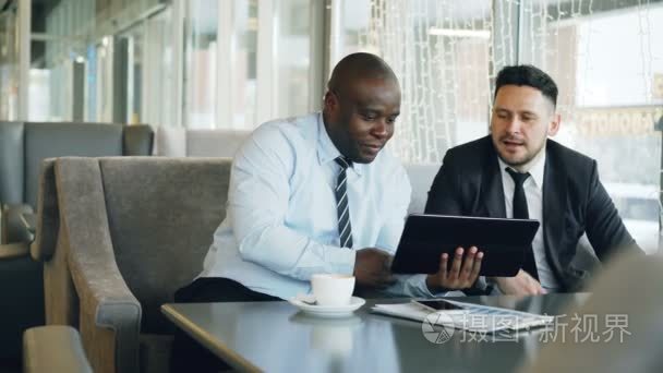 欢快的非洲裔美国商人在正式服装使用数字平板讨论启动项目与他的高加索伙伴在时尚咖啡馆午餐休息时间